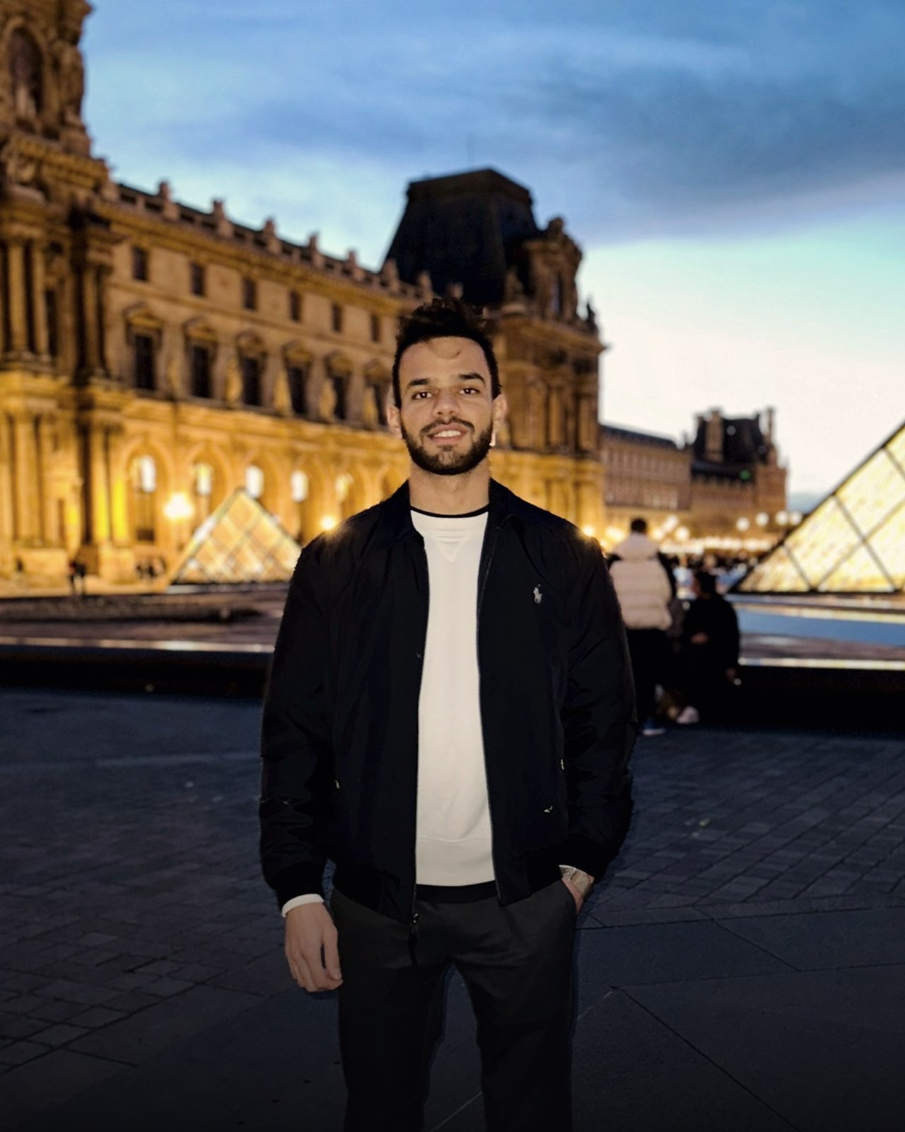 Mateen Rahmat at the Louvre at dusk, the lit pyramid behind him